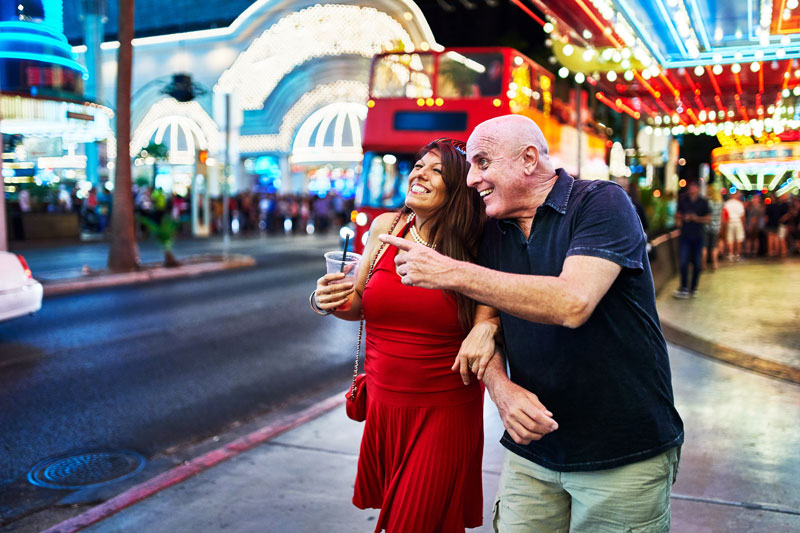 Couple walking along a lively Las Vegas street at night with bright casino lights and entertainment venues in the background.