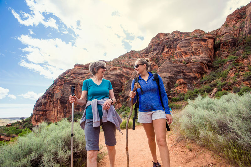 Two women hiking on a desert trail surrounded by red rock cliffs, representing Nevada’s outdoor recreation and scenic landscapes.
